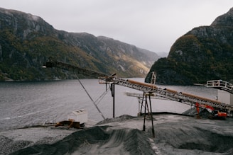 An industrial landscape features a conveyor belt structure set against a backdrop of mountainous terrain and a body of water. The scene includes an expanse of gravel in the foreground, with cloudy skies adding to the dramatic effect.