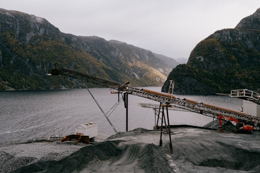 An industrial landscape features a conveyor belt structure set against a backdrop of mountainous terrain and a body of water. The scene includes an expanse of gravel in the foreground, with cloudy skies adding to the dramatic effect.