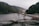 An industrial landscape features a conveyor belt structure set against a backdrop of mountainous terrain and a body of water. The scene includes an expanse of gravel in the foreground, with cloudy skies adding to the dramatic effect.