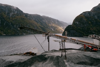 An industrial landscape features a conveyor belt structure set against a backdrop of mountainous terrain and a body of water. The scene includes an expanse of gravel in the foreground, with cloudy skies adding to the dramatic effect.