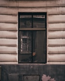 A window is set in a textured building facade with prominent horizontal grooves. The glass of the window shows reflections and appears slightly dirty or weathered. The frame of the window is dark, contrasting with the lighter color of the wall. Subtle reflections and possibly a hint of something inside can be seen on the window's surface.