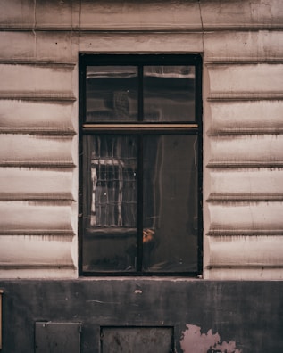 A window is set in a textured building facade with prominent horizontal grooves. The glass of the window shows reflections and appears slightly dirty or weathered. The frame of the window is dark, contrasting with the lighter color of the wall. Subtle reflections and possibly a hint of something inside can be seen on the window's surface.