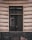 A window is set in a textured building facade with prominent horizontal grooves. The glass of the window shows reflections and appears slightly dirty or weathered. The frame of the window is dark, contrasting with the lighter color of the wall. Subtle reflections and possibly a hint of something inside can be seen on the window's surface.