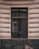 A window is set in a textured building facade with prominent horizontal grooves. The glass of the window shows reflections and appears slightly dirty or weathered. The frame of the window is dark, contrasting with the lighter color of the wall. Subtle reflections and possibly a hint of something inside can be seen on the window's surface.