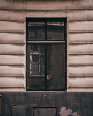 A window is set in a textured building facade with prominent horizontal grooves. The glass of the window shows reflections and appears slightly dirty or weathered. The frame of the window is dark, contrasting with the lighter color of the wall. Subtle reflections and possibly a hint of something inside can be seen on the window's surface.