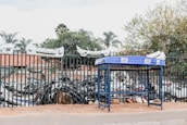 A blue metal stand is positioned on a sidewalk in front of a tall fence. Behind the fence, numerous car bumpers and other automobile parts are stacked together. Several white car bumpers are mounted on top of the fence. Trees and a building with a red roof are visible in the background.