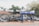 A blue metal stand is positioned on a sidewalk in front of a tall fence. Behind the fence, numerous car bumpers and other automobile parts are stacked together. Several white car bumpers are mounted on top of the fence. Trees and a building with a red roof are visible in the background.