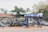 A blue metal stand is positioned on a sidewalk in front of a tall fence. Behind the fence, numerous car bumpers and other automobile parts are stacked together. Several white car bumpers are mounted on top of the fence. Trees and a building with a red roof are visible in the background.
