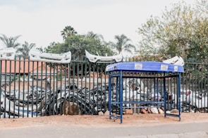 A blue metal stand is positioned on a sidewalk in front of a tall fence. Behind the fence, numerous car bumpers and other automobile parts are stacked together. Several white car bumpers are mounted on top of the fence. Trees and a building with a red roof are visible in the background.