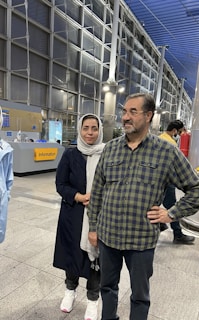 A man and a woman standing together in an indoor public setting, possibly an airport or station. The man is in a plaid shirt and jeans, wearing glasses with a slight smile, while the woman is wearing a dark coat and a light hijab. The background shows a modern architectural design with glass panels, metal structures, and an information sign.