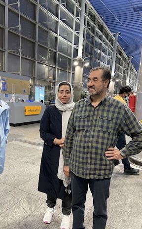 A man and a woman standing together in an indoor public setting, possibly an airport or station. The man is in a plaid shirt and jeans, wearing glasses with a slight smile, while the woman is wearing a dark coat and a light hijab. The background shows a modern architectural design with glass panels, metal structures, and an information sign.