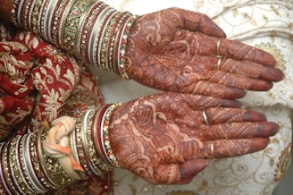 Intricate henna designs adorn the open palms, surrounded by ornate bangles on the wrists. The background features richly embroidered red and gold fabric.