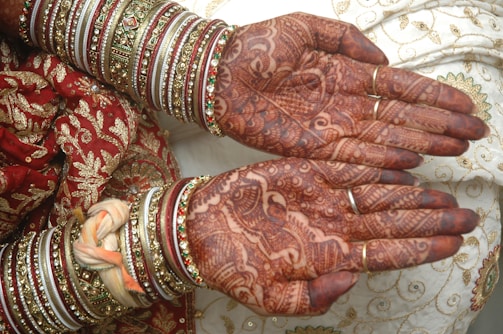 Intricate henna designs adorn the open palms, surrounded by ornate bangles on the wrists. The background features richly embroidered red and gold fabric.