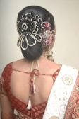 A woman is seen from behind, showcasing an elaborate hairstyle adorned with intricate jewelry and embellishments. She is wearing a traditional outfit with a red patterned blouse and a white saree draped over her shoulder. The hairstyle features a decorative hairpiece with floral and bead designs.