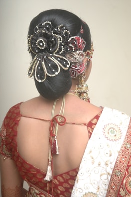 A woman is seen from behind, showcasing an elaborate hairstyle adorned with intricate jewelry and embellishments. She is wearing a traditional outfit with a red patterned blouse and a white saree draped over her shoulder. The hairstyle features a decorative hairpiece with floral and bead designs.