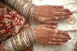 Intricate henna designs adorn the hands, complemented by elaborate bangles in various colors and intricate patterns. The attire features exquisite embroidery with golden and red motifs, highlighting a traditional and festive aesthetic.