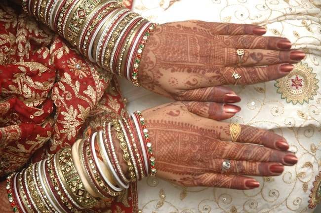 Intricate henna designs adorn the hands, complemented by elaborate bangles in various colors and intricate patterns. The attire features exquisite embroidery with golden and red motifs, highlighting a traditional and festive aesthetic.