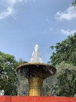 A large water fountain with a golden base and a wide circular brim. Water is cascading over the sides and sprouting upwards, surrounded by lush green trees and under a clear blue sky.