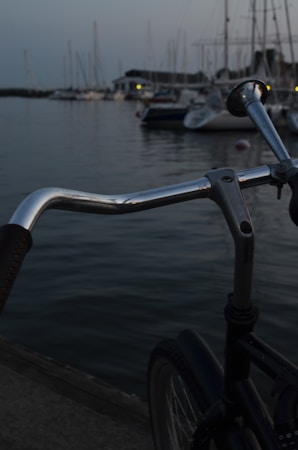 A close-up view of a bicycle handlebar is in the foreground, with a peaceful marina scene in the background. Several boats are docked on calm water, and the scene is dimly lit, suggesting early evening or dusk.