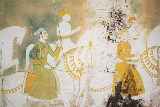 A traditional mural painting depicting figures riding horses. The central figure wears a turban and intricate clothing, holding a spear. The scene is painted with muted earth tones and shows signs of wear and aging, with visible cracks and peeling.