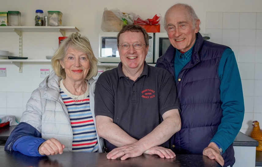 Three people pose together in a kitchen setting, with shelves and kitchen items visible in the background. The individuals are smiling, suggesting a friendly or familial relationship. The central person is wearing a dark shirt with text, while the other two are dressed in casual jackets.