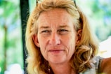 A middle-aged person with long blonde hair gazes outward, wearing sunglasses on their head. The background is a blurred mix of green and blue hues, suggesting an outdoor setting.