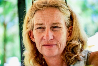 A middle-aged person with long blonde hair gazes outward, wearing sunglasses on their head. The background is a blurred mix of green and blue hues, suggesting an outdoor setting.