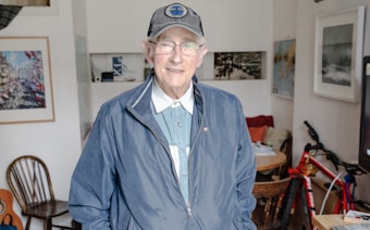 An elderly man wearing a navy jacket and baseball cap stands in a cozy, eclectic living space. The walls are adorned with various pieces of artwork, and there is a bicycle to one side. A table with scattered items and a wooden chair are also visible.