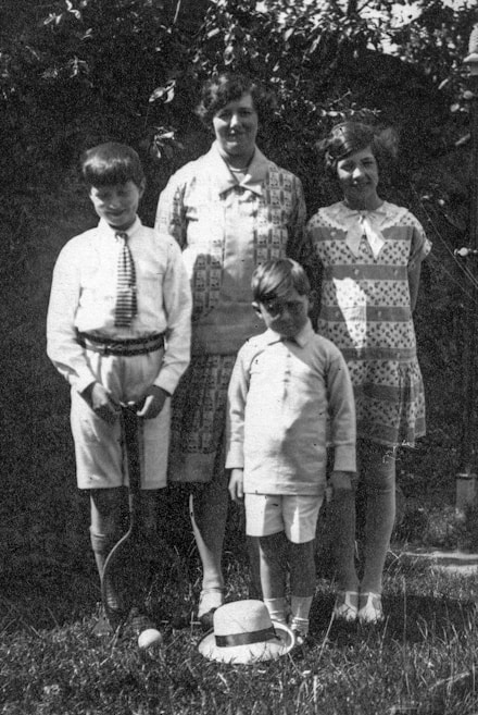 A black and white photograph featuring two women and two boys standing outdoors with trees in the background. The older boy holds a tennis racket and stands next to a tennis ball and straw hat on the grass.