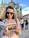 A woman wearing sunglasses and a plaid shirt is reading a travel guidebook in front of a historic Gothic-style cathedral. The cobblestone square and a group of people in the background indicate a popular tourist area.