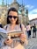 A woman wearing sunglasses and a plaid shirt is reading a travel guidebook in front of a historic Gothic-style cathedral. The cobblestone square and a group of people in the background indicate a popular tourist area.