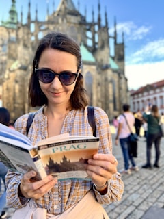 A woman wearing sunglasses and a plaid shirt is reading a travel guidebook in front of a historic Gothic-style cathedral. The cobblestone square and a group of people in the background indicate a popular tourist area.