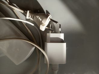 A close-up of two electrical plugs lying on a smooth surface. One plug is attached to a cable, while the other plug adapter is next to it. The image features a combination of shadows and light, highlighting the metallic prongs and white plastic casings.