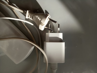 A close-up of two electrical plugs lying on a smooth surface. One plug is attached to a cable, while the other plug adapter is next to it. The image features a combination of shadows and light, highlighting the metallic prongs and white plastic casings.