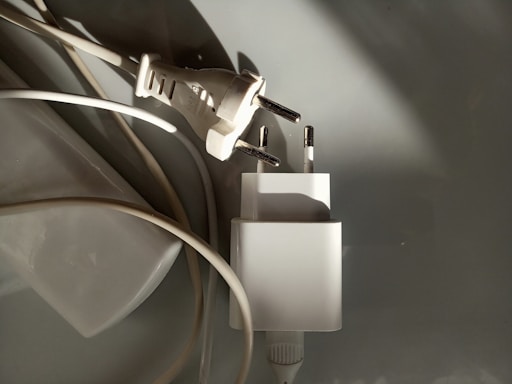 A close-up of two electrical plugs lying on a smooth surface. One plug is attached to a cable, while the other plug adapter is next to it. The image features a combination of shadows and light, highlighting the metallic prongs and white plastic casings.