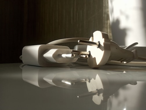 A close-up view of a white electrical plug and adapter lying on a reflective surface. Soft shadows and natural light illuminate the scene, creating a tranquil and calm atmosphere.