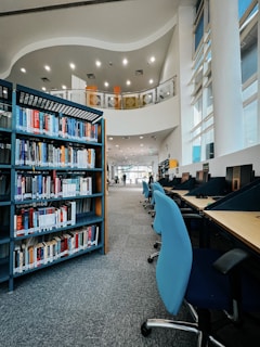 A modern library interior with high ceilings, featuring a long row of blue chairs aligned along desktop computers. There is a tall bookshelf filled with various books, positioned next to the computers. Large windows allow natural light into the space, creating a bright atmosphere. The curved architectural design and minimalist decor add a contemporary touch.
