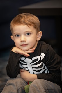 A young child sits with their head resting on one hand, wearing a black shirt with a white skeleton print across the chest. The child has short light brown hair and a gentle expression on their face. The background is softly out of focus, emphasizing the subject.