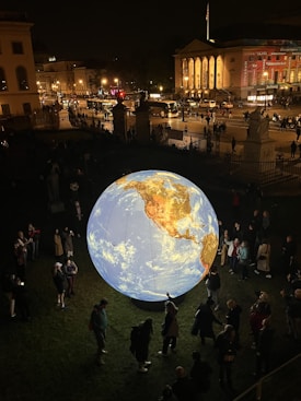 A large, illuminated globe is displayed outdoors at night, surrounded by a crowd of people. The globe features a detailed map with visible continents, notably North and South America. In the background, there are city buildings with bright lights and a few vehicles on the street, creating an urban atmosphere.