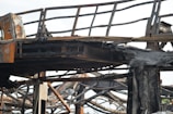 A close-up view of a charred metal structure. The metal beams and framework seem to have been severely damaged by fire. The surface of the metal is blackened and covered with soot, and parts of the structure appear to be twisted and melted.