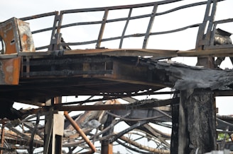 A close-up view of a charred metal structure. The metal beams and framework seem to have been severely damaged by fire. The surface of the metal is blackened and covered with soot, and parts of the structure appear to be twisted and melted.