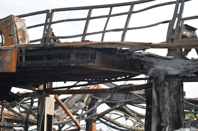 A close-up view of a charred metal structure. The metal beams and framework seem to have been severely damaged by fire. The surface of the metal is blackened and covered with soot, and parts of the structure appear to be twisted and melted.