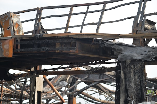 A close-up view of a charred metal structure. The metal beams and framework seem to have been severely damaged by fire. The surface of the metal is blackened and covered with soot, and parts of the structure appear to be twisted and melted.