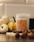 A pumpkin spice scented candle is situated on a surface surrounded by small pumpkins and hazelnuts. The candle has a minimalist label with elegant typography. The warm tones of the objects create a cozy and autumnal atmosphere.