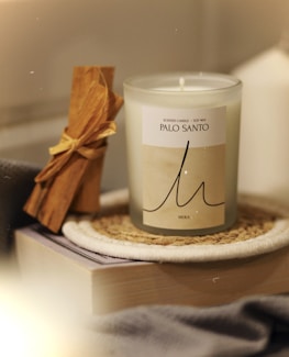 A scented candle labeled 'Palo Santo' in a glass container is placed on a woven coaster. Next to it is a bundle of Palo Santo sticks tied with a yellow ribbon, resting on top of a closed book. The scene exudes a cozy and warm ambiance.