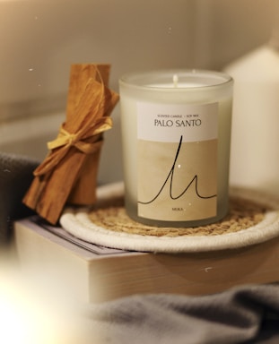 A scented candle labeled 'Palo Santo' in a glass container is placed on a woven coaster. Next to it is a bundle of Palo Santo sticks tied with a yellow ribbon, resting on top of a closed book. The scene exudes a cozy and warm ambiance.