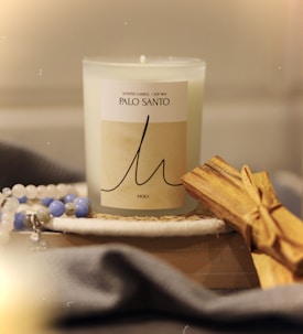A Palo Santo scented candle in a frosted glass container sits on a flat surface. Next to the candle, there is a bundle of Palo Santo sticks tied together. In the foreground, there is a gray, soft fabric, and in the background, there are beaded bracelets with white and blue beads.