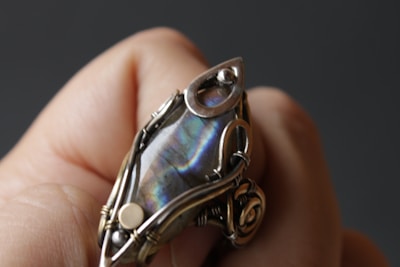 A close-up of a hand holding a handcrafted ring featuring a large, iridescent gemstone. The stone is encased in an intricate wire wrapping design with metallic elements and embellishments.