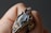 A close-up of a hand holding a handcrafted ring featuring a large, iridescent gemstone. The stone is encased in an intricate wire wrapping design with metallic elements and embellishments.