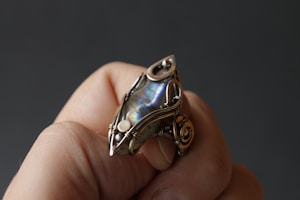 A hand holds an intricately designed ring featuring an iridescent stone set in a detailed metal framework. The metal has an elaborate, wire-like design that spirals around the stone.